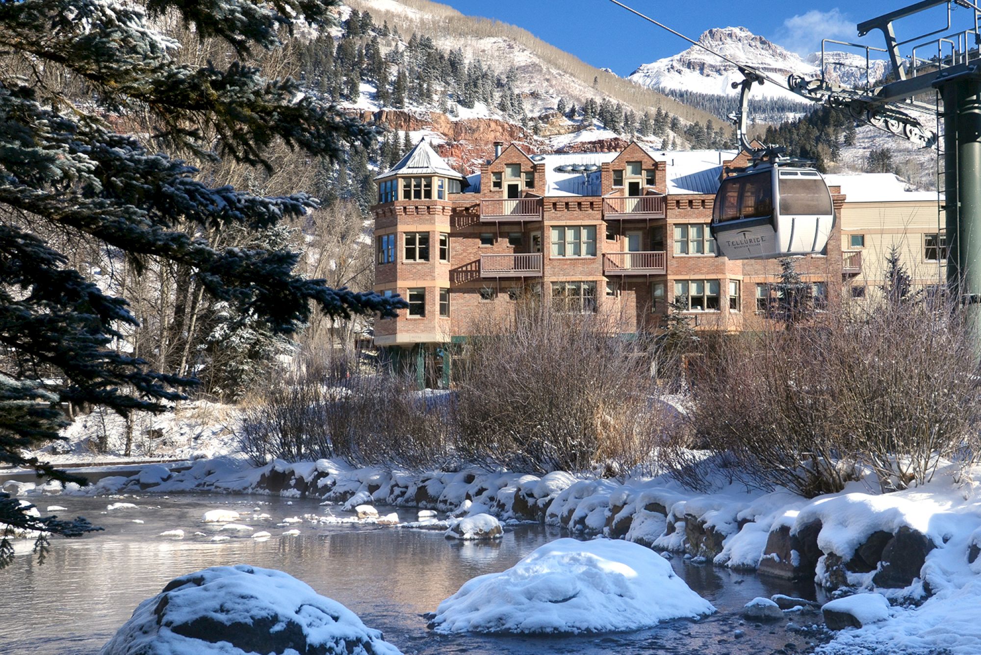 A snowy mountain scene with a building, a gondola lift, and a partially frozen river flowing through the landscape.