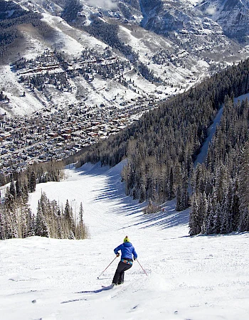 A skier descends a snowy slope with a tree-lined path, overlooking a valley and mountains in the background.