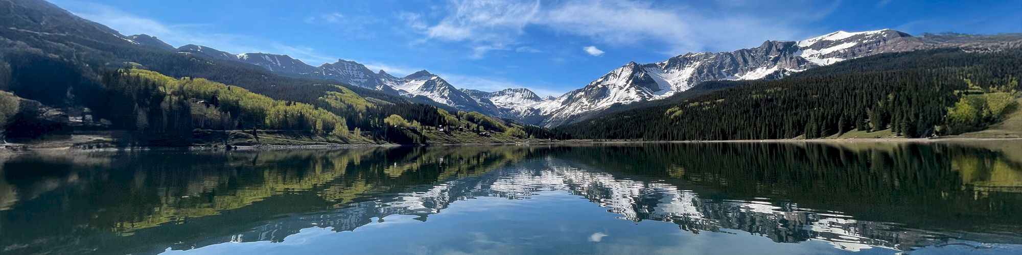 A serene lake reflecting mountains and trees under a sunny sky, with scattered clouds and vibrant greenery.
