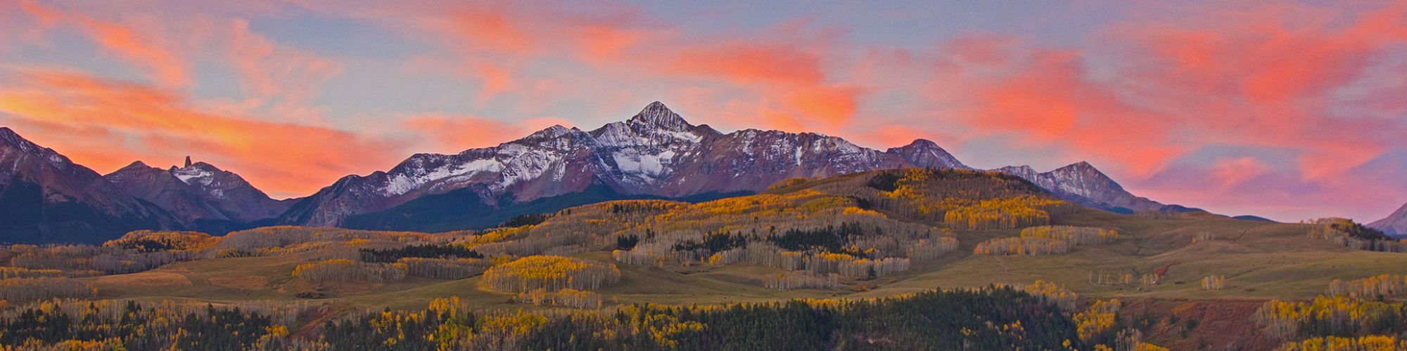 A vibrant sunset casts pink hues over snow-capped mountains and a forested landscape with autumn foliage.