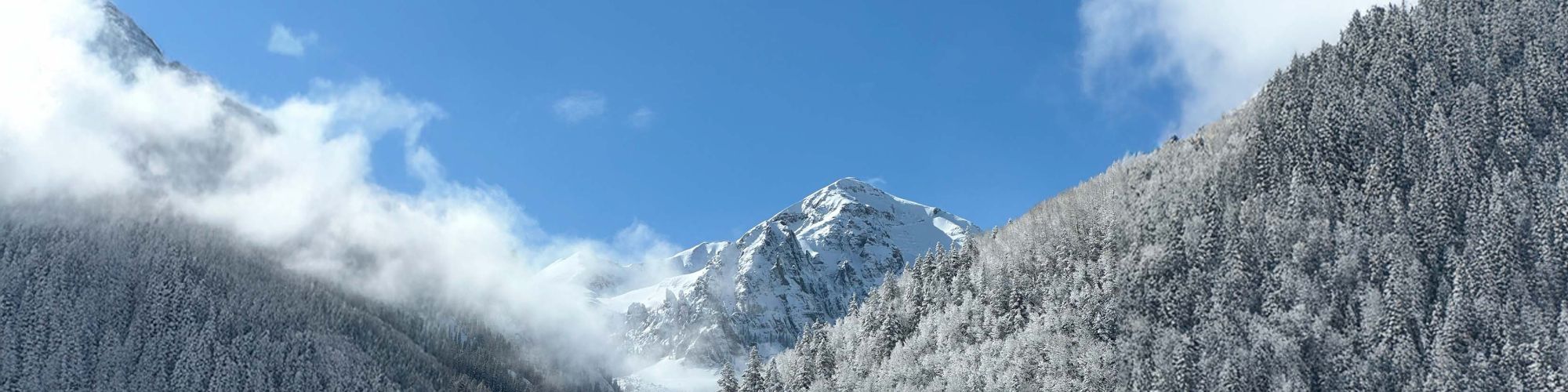 A snowy mountain landscape with clear blue sky, frosted trees, and clouds drifting over the peaks create a serene winter scene.