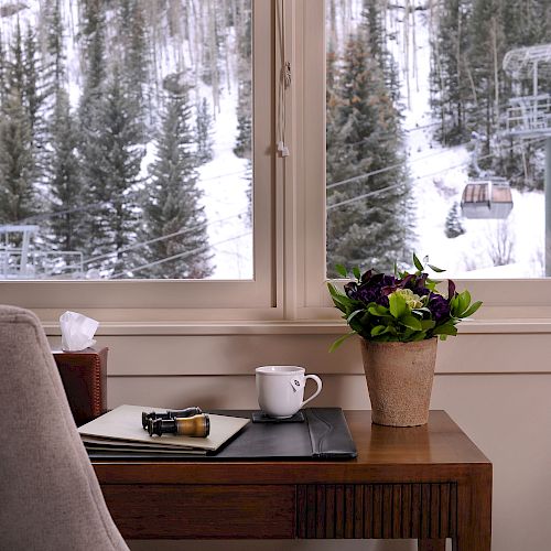 A cozy room with a window view of snowy trees, a desk with a mug and notebook, and a chair.