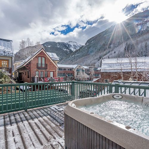 Hot tub on a snowy deck with mountain view and houses. Sun peeks through clouds.