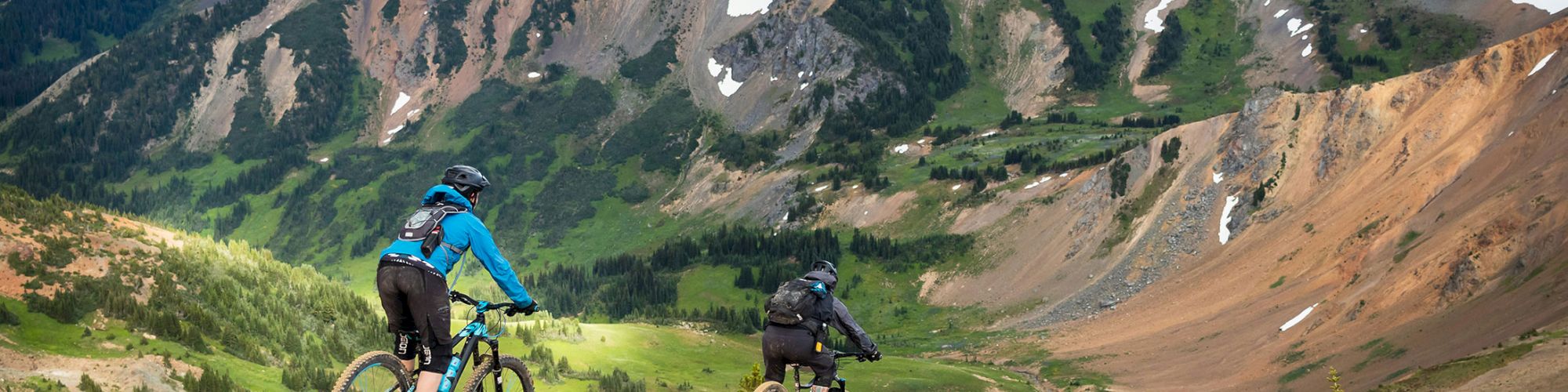 Two people are mountain biking on a trail with picturesque mountains and valleys in the background, under a cloudy sky.