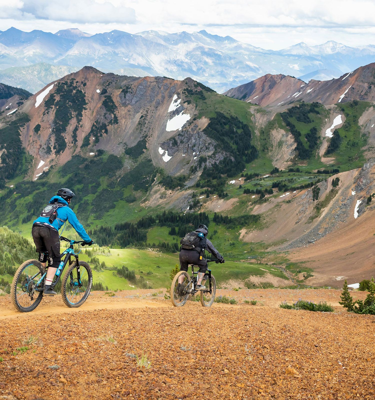 Two cyclists ride mountain bikes down a dirt trail, surrounded by scenic mountains with patches of snow and greenery, under a partly cloudy sky.