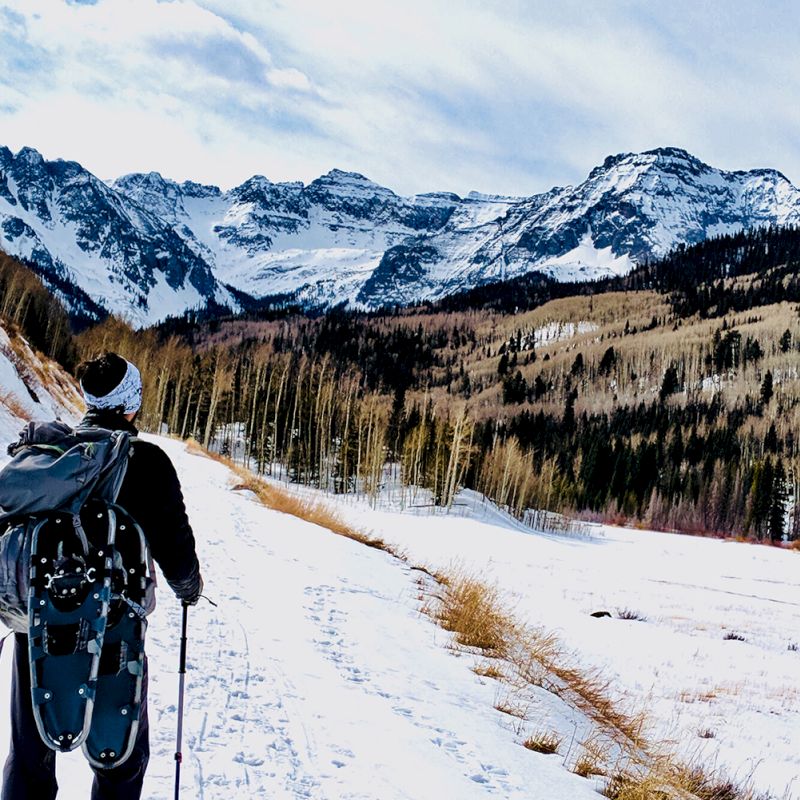 A person with a backpack and snowshoes stands on a snowy path, surrounded by mountains and trees.