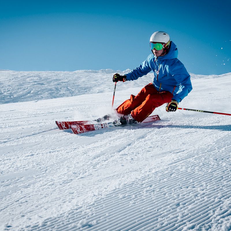 A skier in a blue jacket and red pants descends a snowy slope under a clear sky, kicking up snow as they turn.