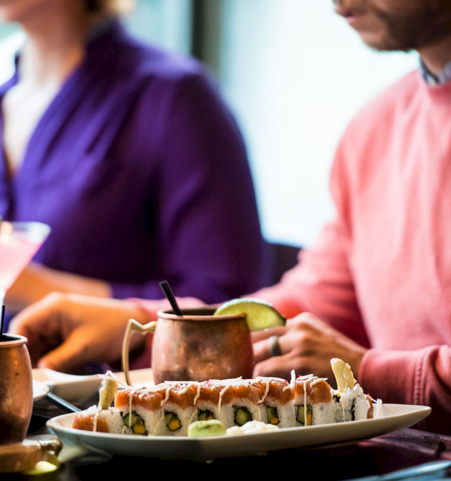 Two individuals are dining with sushi rolls, a cocktail, and two copper mugs with lime garnishes in a restaurant setting.