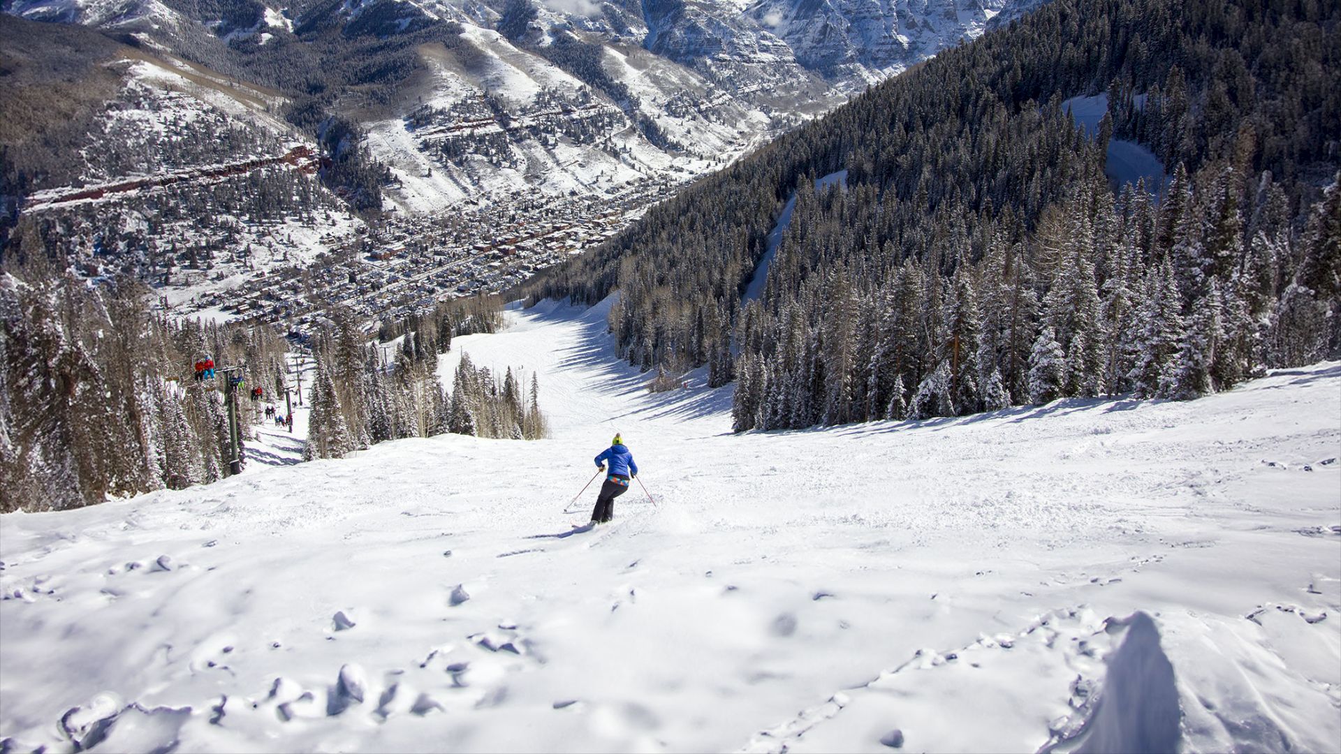 A person skiing down a snowy slope surrounded by pine trees, with a mountainous landscape and a town in the valley below under a blue sky.