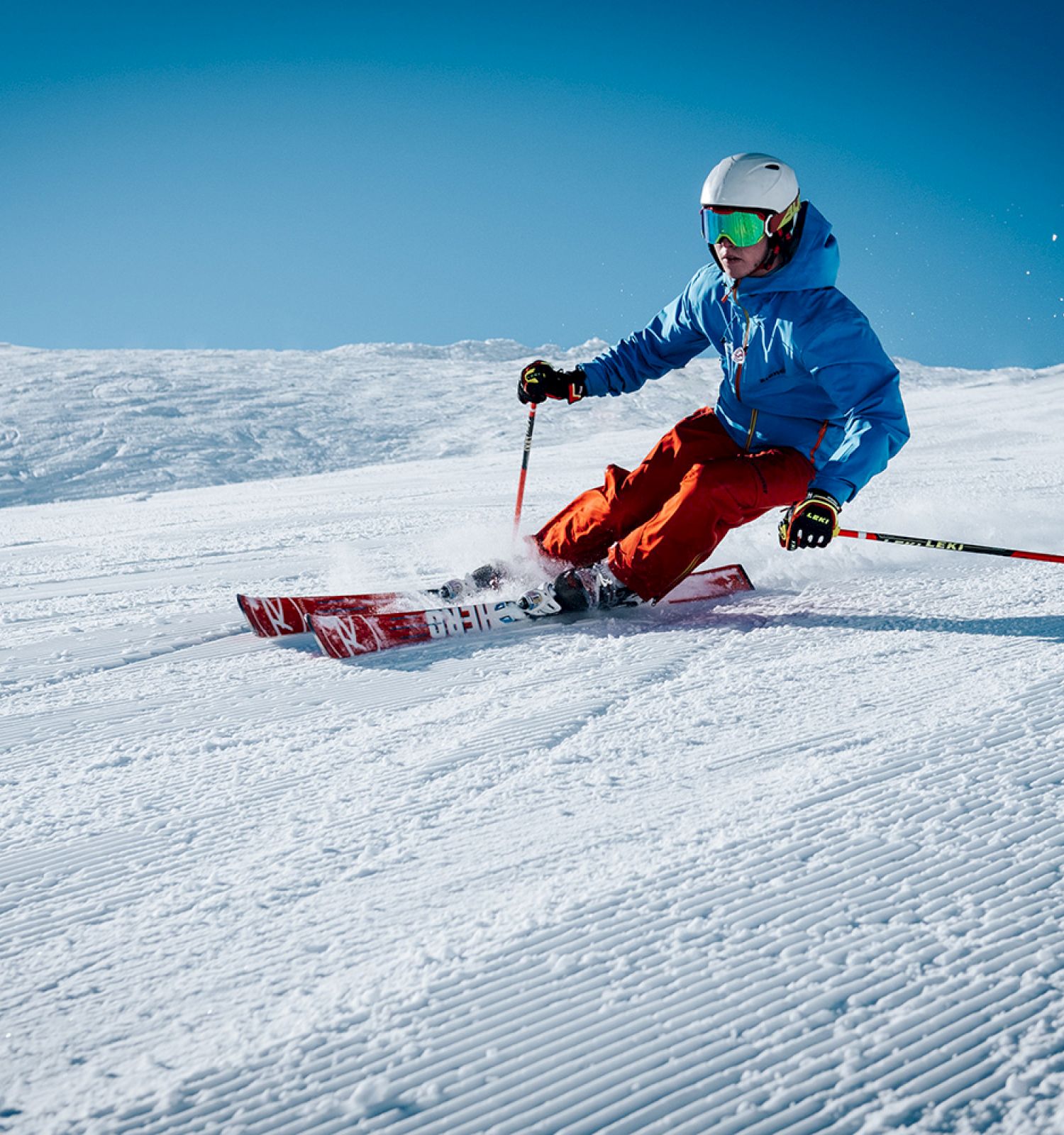 A person skiing down a snowy slope, wearing a blue jacket, red pants, and a white helmet, against a clear blue sky.