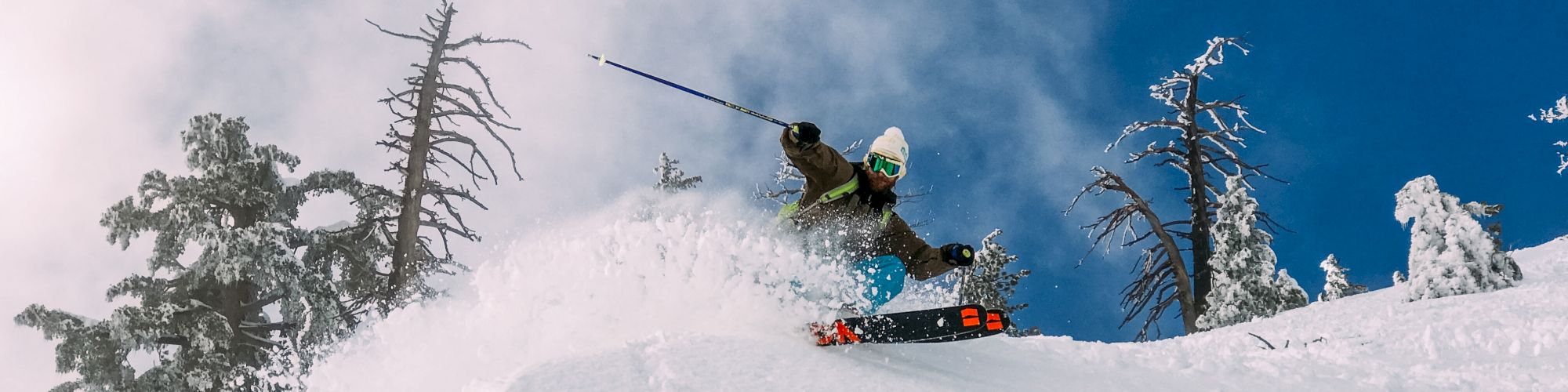 A skier is descending a snowy slope, kicking up powder in a snowy landscape with a bright blue sky.