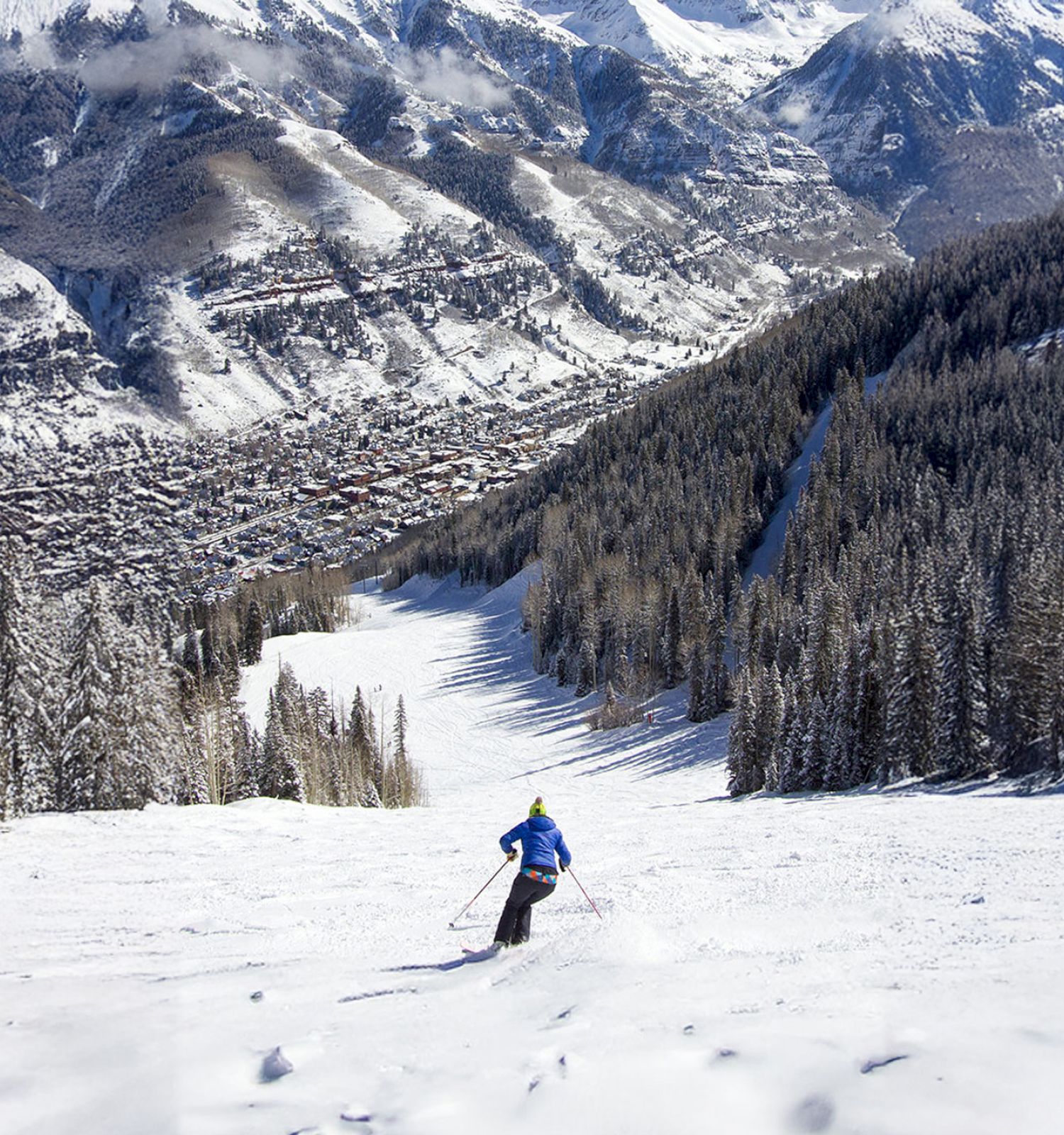 A skier descends a snowy slope surrounded by pine trees and a mountainous landscape, with a town visible in the valley below.