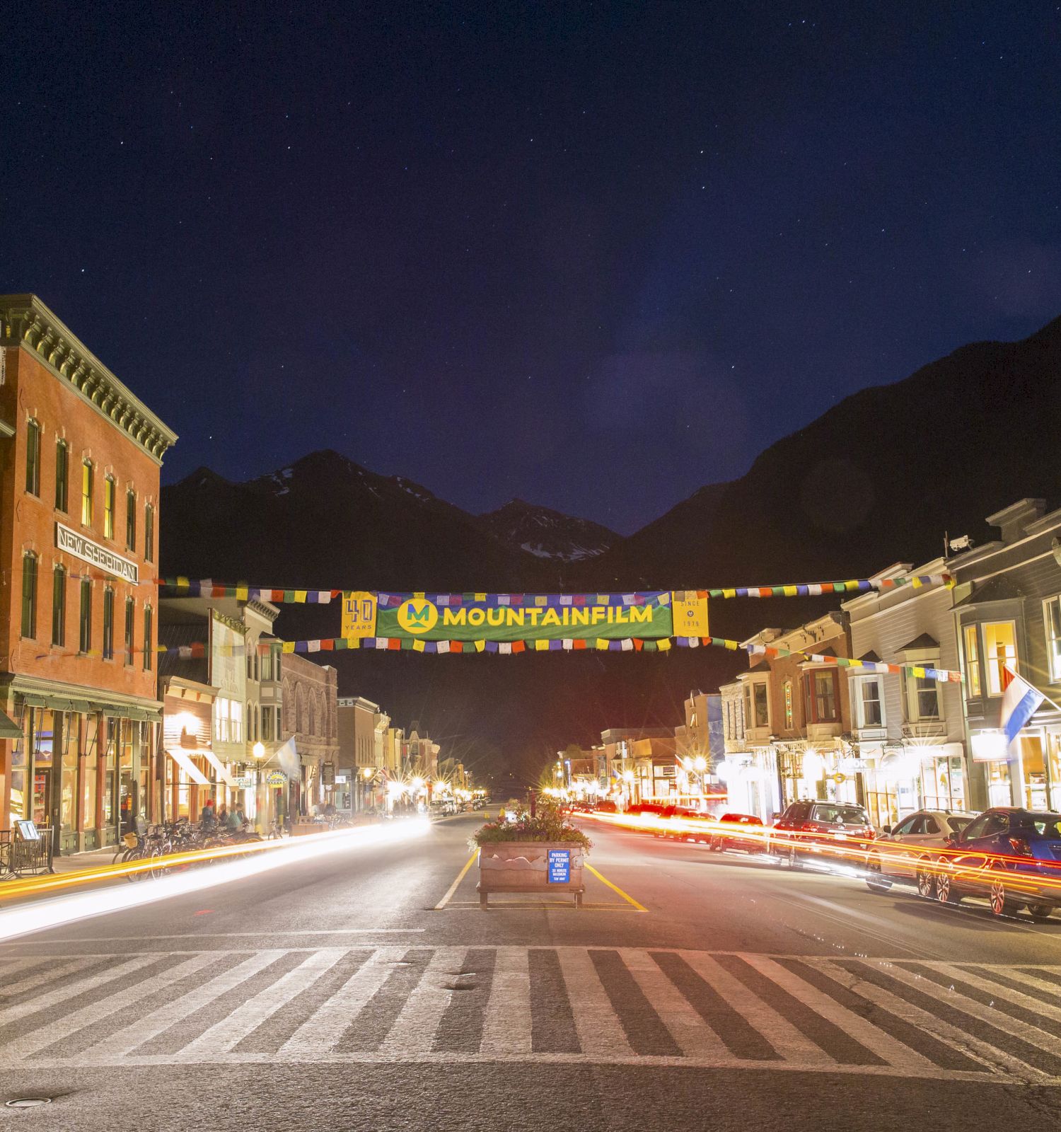 A nighttime street scene with buildings, a festival banner, and mountains in the background, illuminated by streetlights and car headlights.