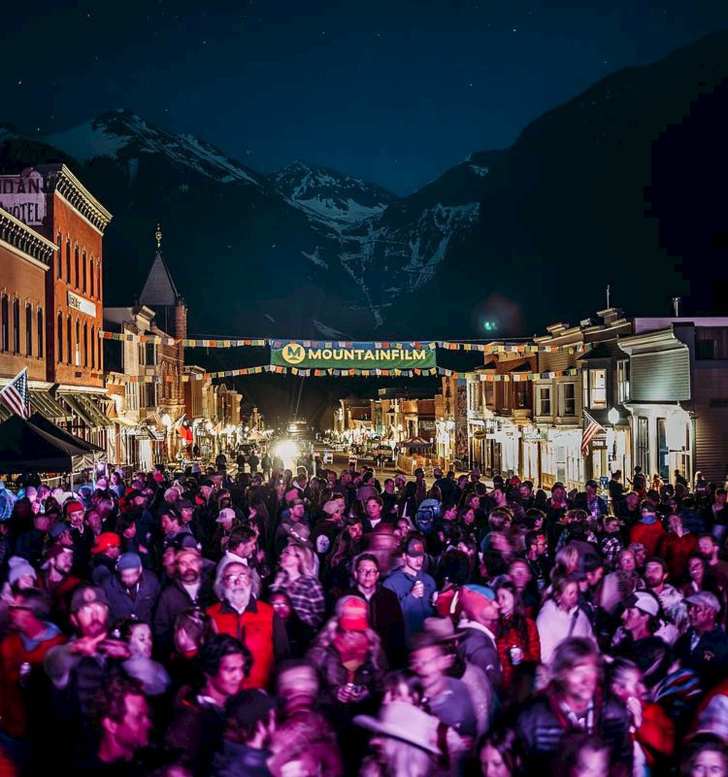 A large crowd gathers outdoors at night in a town with snowy mountains in the background, possibly for an event or festival.