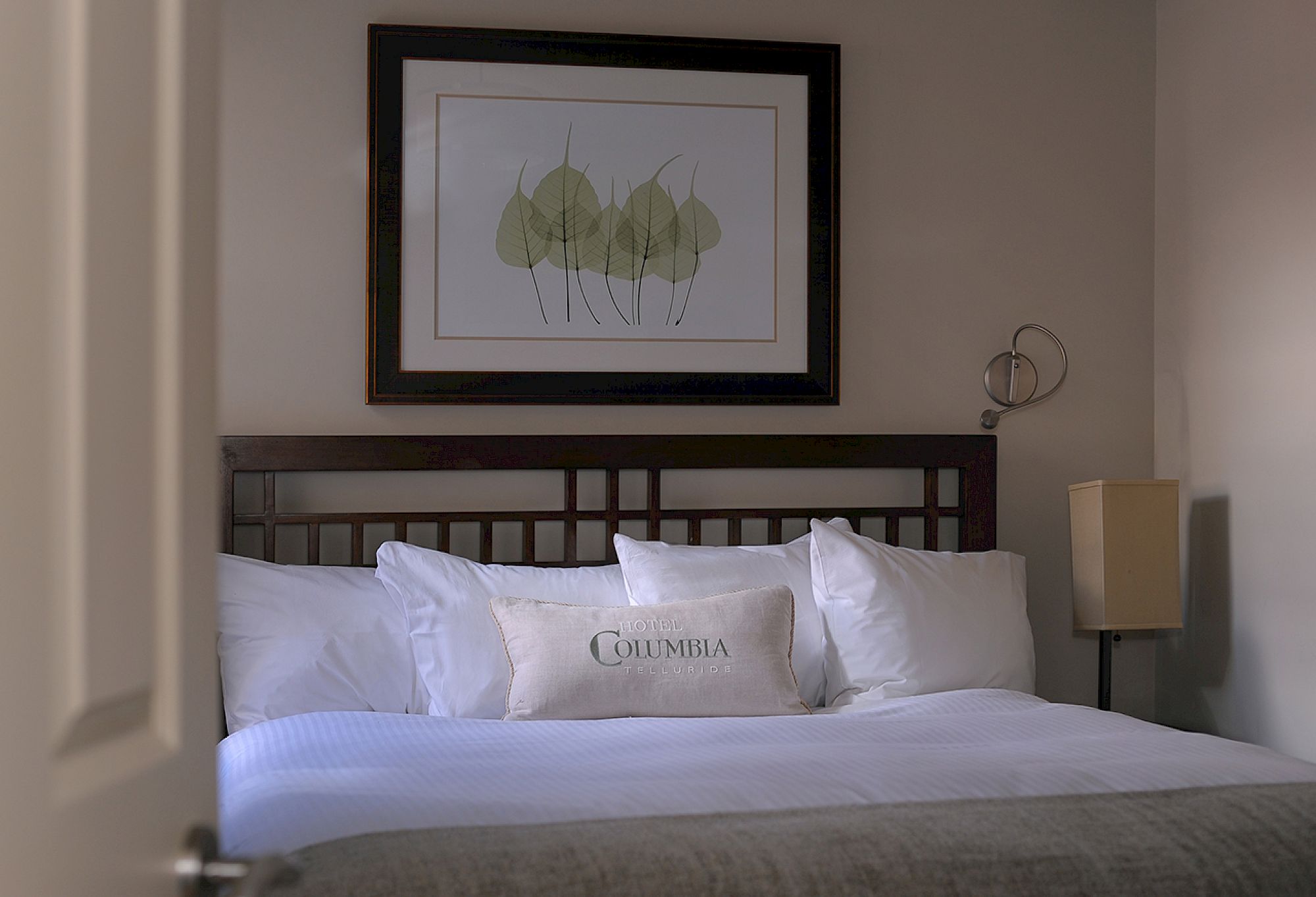 A neatly made bed with white pillows, a framed leaf artwork above, and a lamp to the side, viewed through a partially open door.