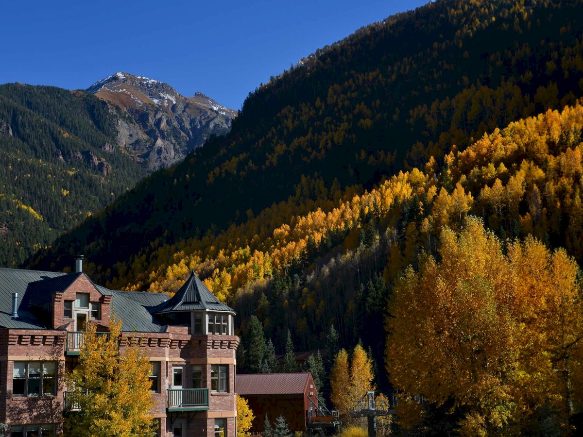 A scenic autumn landscape features a building amid golden trees, set against a backdrop of forested mountains and a clear blue sky.