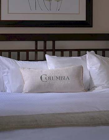 A bed with white pillows, including one with "COLUMBIA" on it, and a bed frame beneath a picture of abstract trees on the wall.